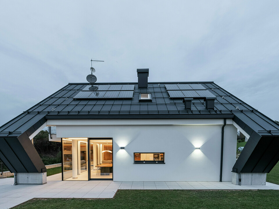Modernes Einfamilienhaus in Borgo Valbelluna, Italien, mit markantem Satteldach aus PREFALZ in P.10 Anthrazit. Die langlebige Aluminium-Dacheindeckung bietet optimalen Schutz vor Witterungseinflüssen und überzeugt durch flexible Verlegemöglichkeiten. Integrierte Photovoltaikmodule nutzen die Dachfläche effizient zur nachhaltigen Energiegewinnung. Die Kombination aus anthrazitfarbenem Aluminiumdach und weißer Fassade sowie die markante Auskragung des Satteldaches unterstreichen das elegante und zeitlose Design des Gebäudes.