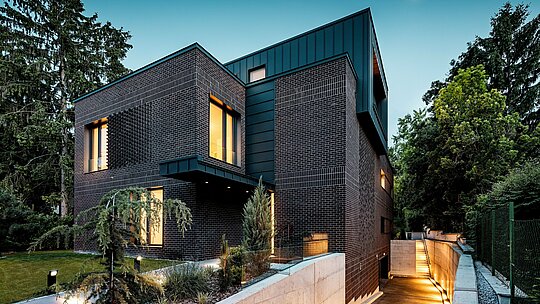 Fotografija prikazuje sodoben, jasno strukturiran zunanji videz večgeneracijske vile v Budimpešti v večernem vzdušju. Temno rdečkastorjave fasade iz opeke se združujejo s precizno izvedenimi PREFALZ aluminijastimi oblogami v barvi P.10 antracit, ki s svojimi navpičnimi linijami ustvarjajo močan arhitekturni poudarek. Toplo osvetljeni okenski odprtini stavbi dodajata globino in atmosfero. Nežno osvetljen dovoz do podzemne garaže elegantno vodi ob stavbi in poudarja visokokakovostno, trajno in vremensko odporno kombinacijo materialov iz klinkerja in PREFA aluminija.