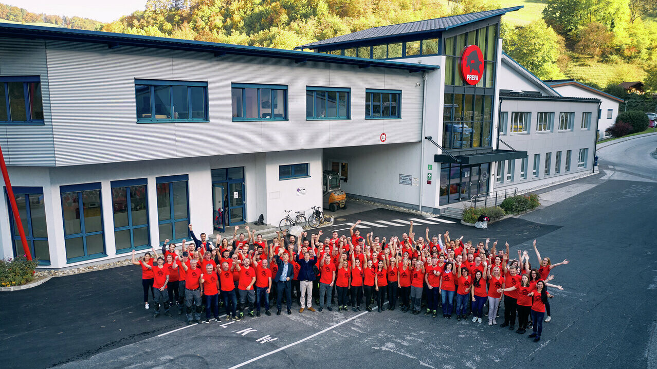 PREFA MitarbeiterInnen mit rotem PREFA T-Shirt stehen in einem Halbkreis vor dem Bürogebäude in Marktl.