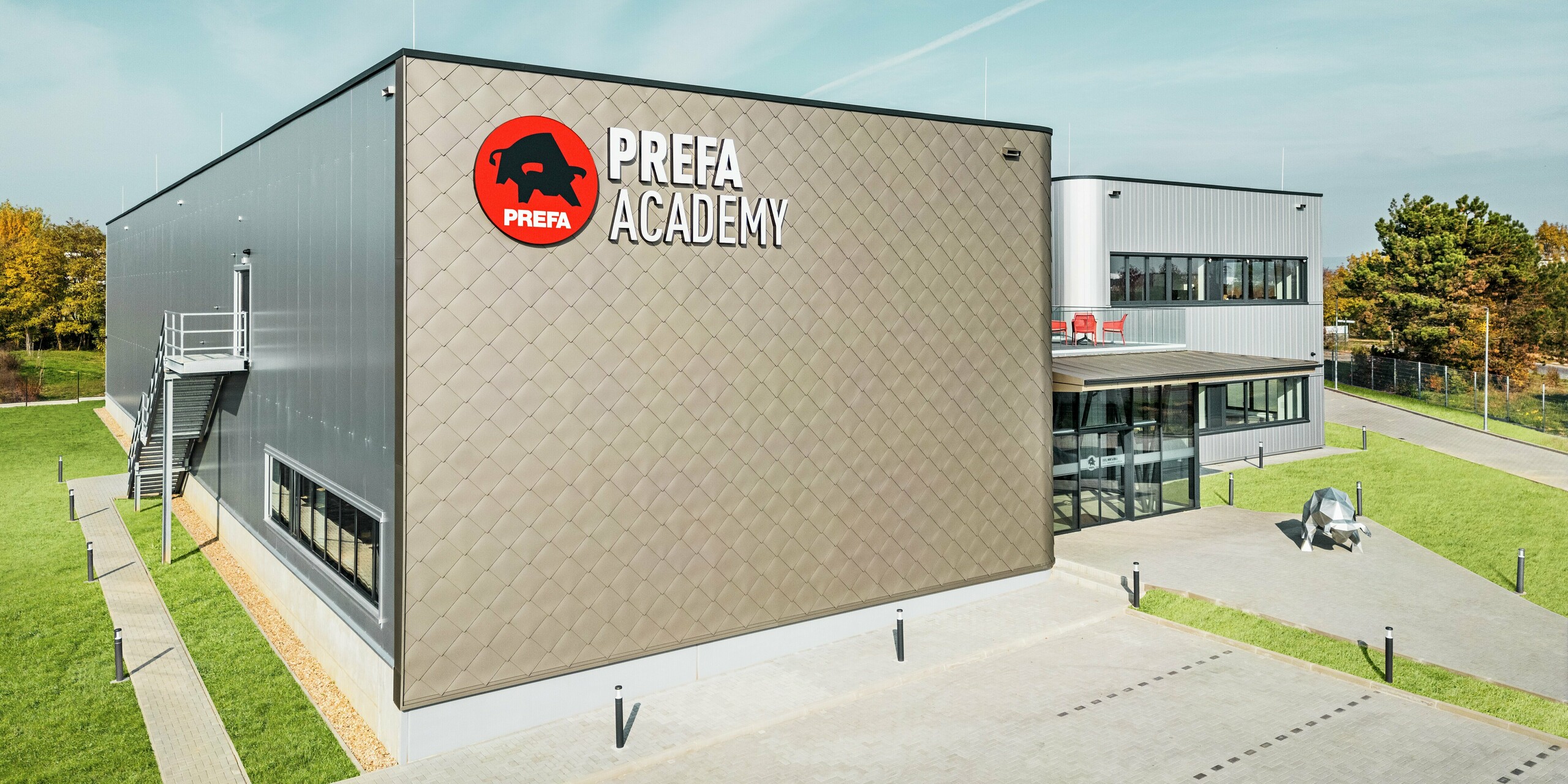 PREFA akademija v Törökbálintu se predstavlja z izrazito fasado iz aluminijastih rombov PREFA 44 × 44 v bronasti barvi in fasadnimi elementi PREFA Siding v srebrno kovinski barvi. Ti visokokakovostni aluminijasti izdelki zagotavljajo sodoben videz, izjemno odpornost na vremenske vplive in dolgo življenjsko dobo. Velik logotip PREFA na fasadi proti parkirišču ter skulptura bika ob poti do vhoda poudarjata moč blagovne znamke.