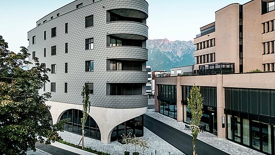 Pogled na Bernerhaus v Buchsu z značilno trikotno obliko in nežno zaobljenimi balkoni pred alpskim gorskim ozadjem. Neprekinjena fasada iz PREFA fasadnih skodel v barvi P.10 svetlo siva obdaja stavbo kot luskasta aluminijasta koža in ustvarja živahno igro svetlobe in sence. Nad masivnim betonskim podstavkom z elegantnimi arkadami deluje lahka, vremensko odporna PREFA aluminijasta fasada še posebej natančna, trajna in enostavna za vzdrževanje – arhitekturni magnet za poglede v urbanem okolju. 