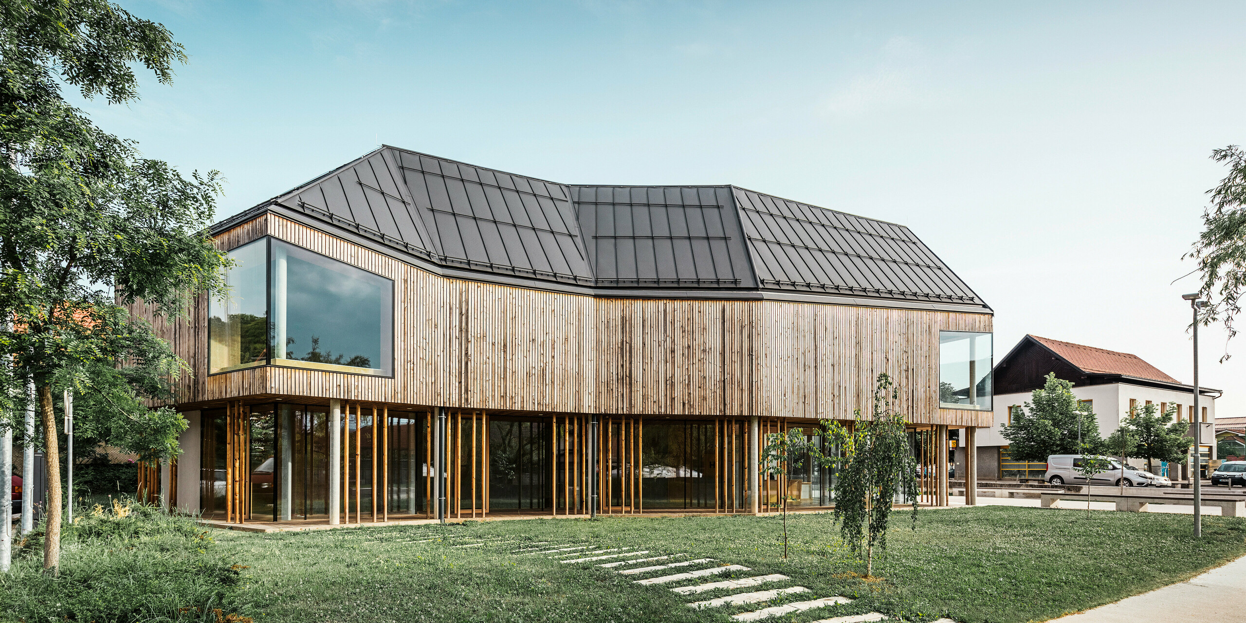 Pogled s strani na ekološko zasnovan center za obiskovalce na Igu v Sloveniji s streho PREFALZ v P.10 temno sivi barvi, ki se harmonično zliva s podeželskim okoljem. Navpična lesena obloga in obsežna zasteklitev poudarjata odprto in pregledno arhitekturo, medtem ko okoliška vegetacija in jasno nebo poudarjata naravno estetiko lokacije. Ta objekt ponazarja prispevek podjetja PREFA Aluminiumprodukte GmbH k sodobni in okolju prijazni arhitekturi, kot je prikazano v knjigi PREFARENZEN 2023.