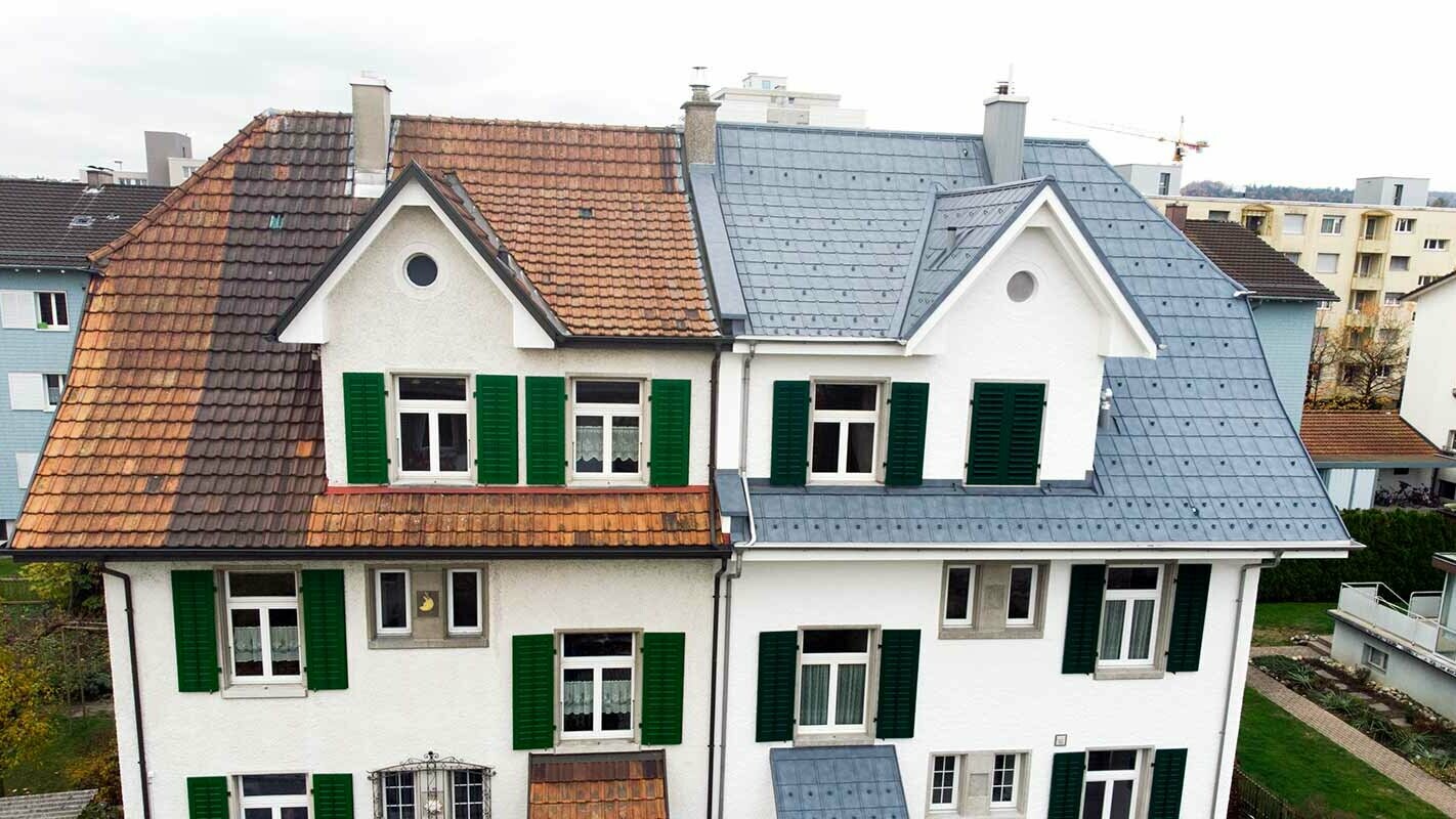 Polovica dvostanovanjske hiše v Švici – levi del zgradbe še ni saniran in je še pokrit z umazanimi strešniki. Desni del zgradbe je saniran s strešnimi ploščami PREFA v kamnito sivi barvi.